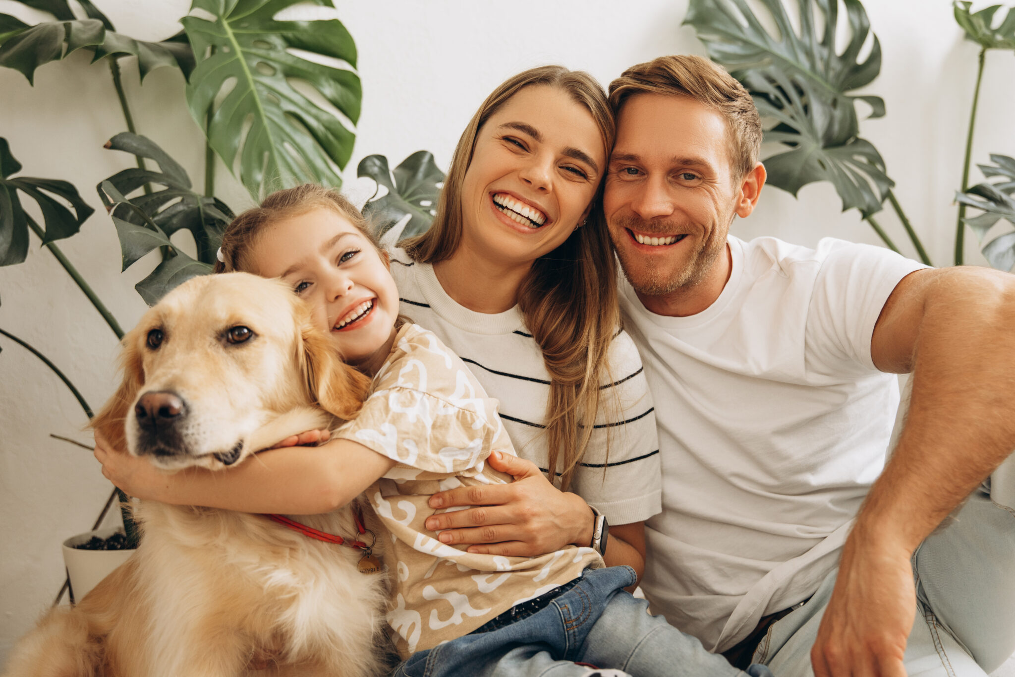 Smiling family with young child and golden retriever sitting indoors, surrounded by houseplants